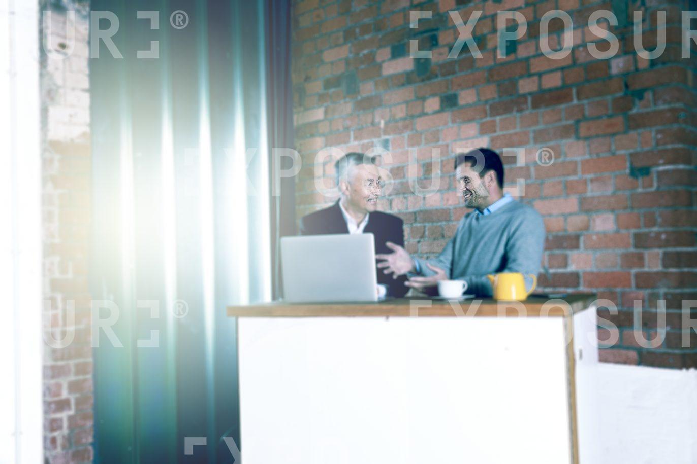 Two Men Stood Up Chatting Around Laptop In Rustic Office